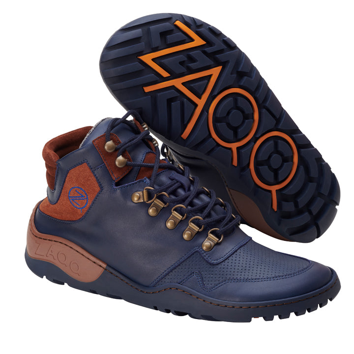 Ein Paar stylische, marineblaue High-Top-Stiefel mit braunen Akzenten, robusten dunklen Sohlen und aufwendigen Schnürdetails. Das wasserdichte Design ZAQQ VAQATION Cognac zeigt orange und braune geometrische Muster auf der Sohle. Diese Outdoor-Schuhe stehen einmal aufrecht und einmal auf der Seite.