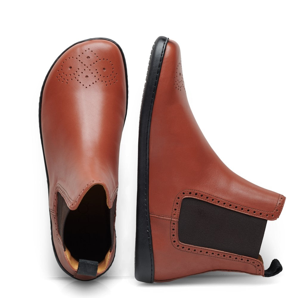 Ein Paar EQUITY Brogue Cognac-Stiefel von ZAQQ besteht aus braunem Leder im Chelsea-Stil und ist mit dekorativen Perforationen an den Zehen verziert. Der linke Stiefel wird aufrecht präsentiert, wodurch die Aufreißlasche hervorgehoben wird, während der rechte Stiefel auf der Seite liegt, um den schwarzen elastischen Zwickel hervorzuheben.