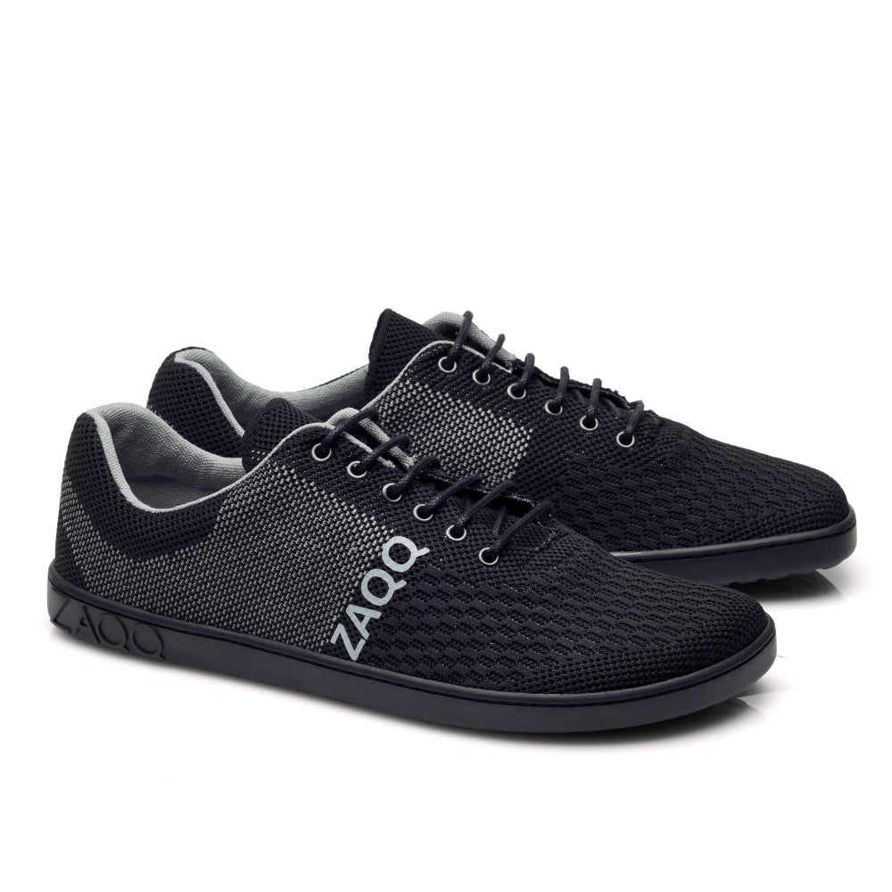 Die QNIT Black Sneaker von ZAQQ sind ein Paar schwarz-graue Mesh-Schuhe mit strukturierter Oberfläche und schwarzen Schnürsenkeln. Diese veganen Strickschuhe tragen das ZAQQ-Logo in Weiß auf der Seite und haben flache, schwarze Sohlen. Sie sind aus atmungsaktivem Baumwollgewebe für mehr Komfort gefertigt.